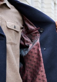 Abrigo de lana azul marino con un forro de patrón burdeos, que presenta una textura suave y un bolsillo oculto, complementando una camisa de botonadura en color beige debajo.