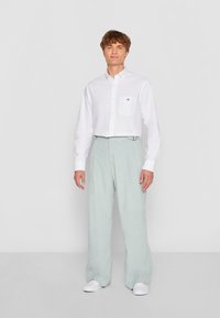 Weißes Hemd mit Knopfleiste und Tasche, kombiniert mit hellgrünen, plissierten, weiten Hosen. Modell trägt weiße Sneakers und steht vor einem schlichten Hintergrund.