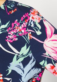 Blusa floral com fundo azul-marinho. O design inclui flores vibrantes em tons de rosa, vermelho e azul, com folhas verdes, feita de um tecido leve.