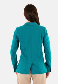 Blazer teal avec un design ajusté, une fente simple à l'arrière et des couturestailorées. Il a des manches longues et un col classique.