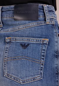 Detailní záběr zadní kapsy modrých džín s logem Emporio Armani a kovovým emblémem orla nad kapsou.