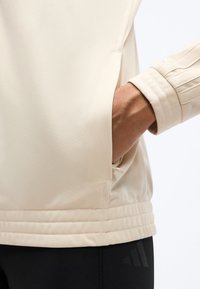 Χέρι στην τσέπη ενός μπεζ σακακιού με ριμπ μανσέτα, εμφανές ράψιμο, φορεμένο πάνω από μαύρο παντελόνι με αχνό λογότυπο Adidas.