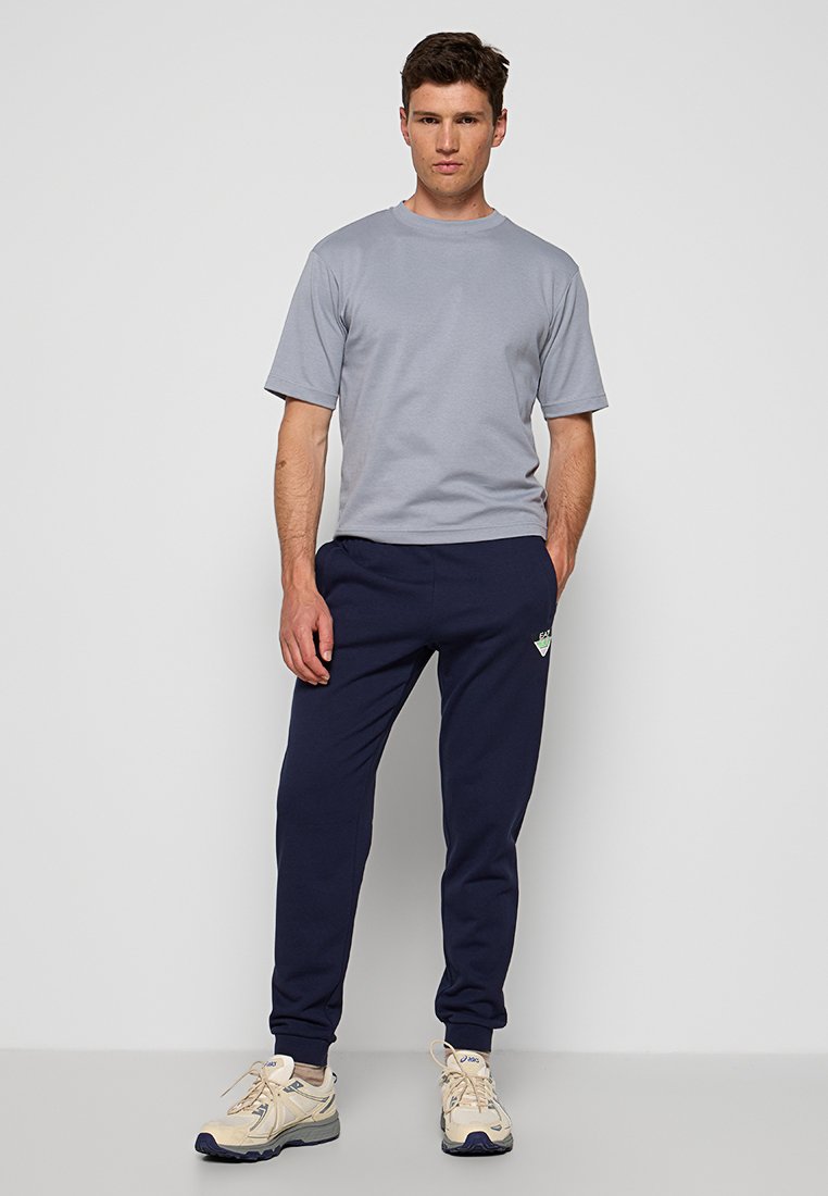 Szara koszulka z krótkim rękawem, granatowe spodnie dresowe z bocznymi kieszeniami, akcent z logo, noszone z beżowymi trampkami. Prosty, luźny strój sportowy.