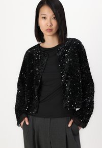 Giacca nera con paillettes, dalla texture morbida, colletto rotondo e design corto. Presenta polsini trasparenti neri e una parte anteriore senza bottoni.