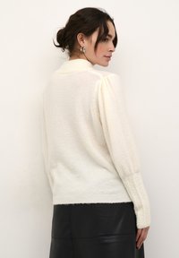 Pull en maille couleur crème avec un col montant, des manches bouffantes et un ourlet texturé. Porté avec une jupe en cuir noire pour le contraste.