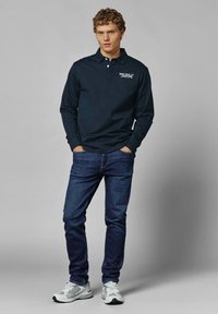 Mornarsko modra polo majica z dolgimi rokavi, ovratnikom in logotipom, v kombinaciji z modrimi kavbojkami in sivi športni čevlji. Enobarvni odtenki, sproščen dizajn in prilega silhueta.