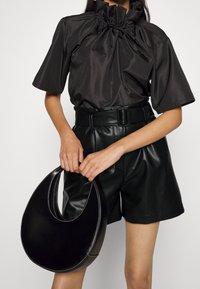 Blouse en satin noir avec un col froncé, manches courtes ; associée à un short en cuir noir et un sac à main courbé noir avec des coutures blanches.