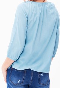 Blusa azzurro chiaro con maniche a tre quarti, texture morbida, scollatura arricciata e vestibilità rilassata, abbinata a jeans scuri strappati.
