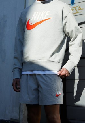 Mężczyzna ubrany w szarą bluzę Nike z czerwonym logo i pasujące szorty stoi przy jasnej ścianie, poprawiając rąbek bluzy.