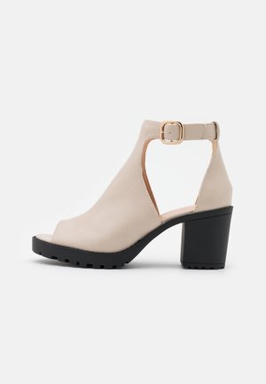 Beige åben tå sandaler med hæl, sort chunky sål, ankelstrop og guld spænde, sidevisning på hvid baggrund.