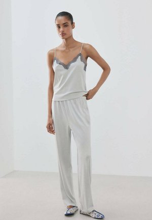 Femme portant un caraco en satin gris clair avec bordure en dentelle et un pantalon large assorti, debout dans une pièce blanche minimaliste.