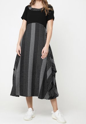 Vrouw die een zwart-wit geruite midi-jurk met korte mouwen en witte sneakers draagt, staand tegen een effen achtergrond.