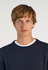 Marineblauer Strickpullover mit strukturiertem Muster, Rundhalsausschnitt und einem weißen T-Shirt darunter. Schlichtes Design und glatte Stofftextur.