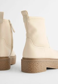 Beige knöchelhigh Stiefel aus einem glatten synthetischen Material mit einer strukturierten Plateausohle und einem Zugband. Dezente Nähte überall.