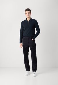 Marineblauwe rits-polo trui, gecombineerd met zwarte corduroy broek met textuurknie-patches en witte sneakers, op een effen achtergrond.