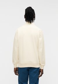 Μπλούζα φούτερ σε κρεμ χρώμα με ψηλή λαιμόκοψη, ριμπ μανσέτες και άνετη γραμμή, συνδυασμένη με μπλε παντελόνι. Μαλακό βαμβακερό ύφασμα. Οπίσθια όψη.