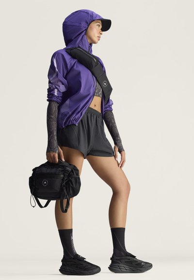 Frau in sportlicher Kleidung mit violettem Kapuzenjacke, schwarzen Shorts und Turnschuhen, die eine schwarze Tasche hält und eine Umhängetasche trägt.