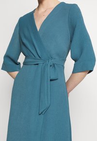 Robe en teal avec un décolleté en V profond, des manches trois-quarts et une taille nouée. Tissu texturé avec un détail de côtes subtil.