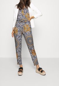 Combinaison à motifs dans des tons de jaune, noir et gris. Dotée d'une taille ajustable et serrée à la taille, associée à un blazer blanc.