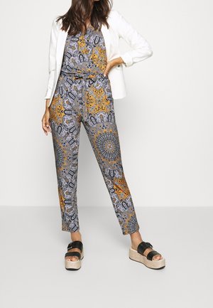 Jumpsuit met patroon in tinten geel, zwart en grijs. Voorzien van een verstelbare taille, gesnoerd in de taille, gecombineerd met een wit colbert.