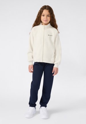 Meisje met lange, krullende haren in een off-white, ritssluitingjasje, marineblauwe joggingbroek en witte sneakers, staande tegen een effen achtergrond.