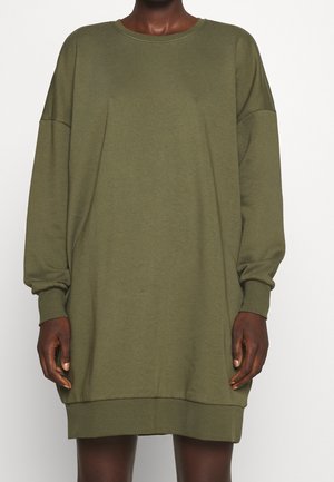 Person iført en løs, langærmet, olivengrøn sweatshirt-kjole med ribkanter ved ærmer og kant, stående mod en ensfarvet baggrund.