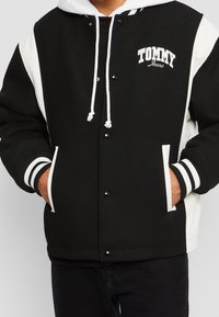 Černobílá varsity bunda s patentkami, žebrovanými manžetami a kapucí. Na hrudi má logo "TOMMY Jeans" a kontrastní design rukávů.