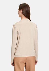 Beige langärmliges Oberteil mit weicher Textur, das mit gerafften Details an den Schultern und einem runden Ausschnitt ausgestattet ist, kombiniert mit hellen Hosen.