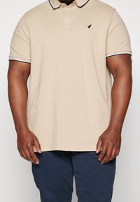 Beżowa koszulka polo z krótkimi rękawami, kołnierzykiem i granatowymi mankietami. Na lewej piersi małe czarne logo. Materiał bawełniany, luźny krój.