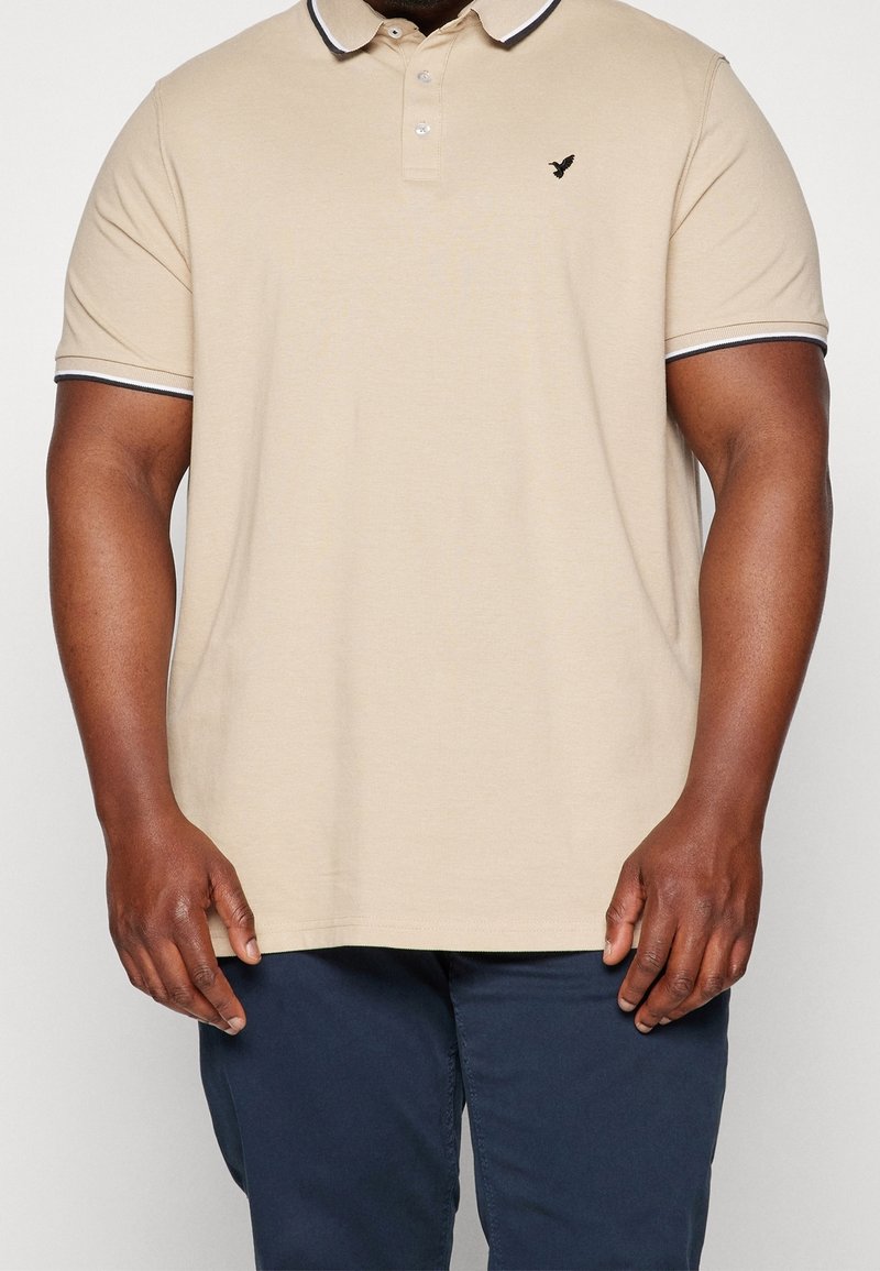 Beżowa koszulka polo z krótkimi rękawami, kołnierzykiem i granatowymi mankietami. Na lewej piersi małe czarne logo. Materiał bawełniany, luźny krój.