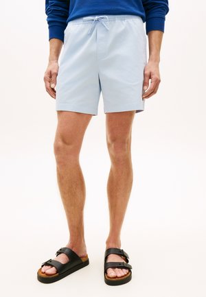 Mężczyzna w jasnoniebieskich szortach ze sznurkiem, czarnych sandałach na klamrę i niebieskiej koszuli z długim rękawem, stojący na tle jednolitego białego tła.