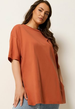 Kobieta z długimi włosami ubrana w oversize'owy, paląco pomarańczowy T-shirt i jasnoniebieskie jeansy, pozująca na jednolitym jasnym tle.