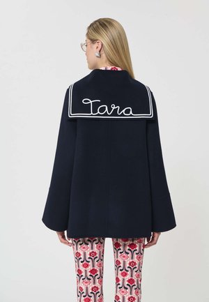 Mujer con cabello rubio lacio que lleva un abrigo azul marino con ribetes blancos y "Tara" bordado en la parte trasera, junto con pantalones de estampado floral.