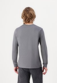 Person mit mittellangem, welligem Haar, die ein einfaches graues Langarmshirt und dunkle Hosen trägt, schaut mit dem Rücken zum Betrachter vor einem weißen Hintergrund.