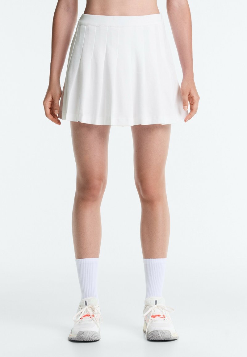 Femme portant une jupe de tennis plissée blanche, des chaussettes blanches mi-mollet et des chaussures de sport blanches, debout contre un fond clair uni.