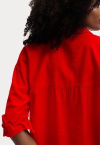 Camicia rossa con un fit rilassato, maniche arrotolabili, dettaglio della piega posteriore e un texture morbida. Il tessuto è leggero e leggermente trasparente.