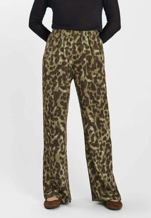 Persoon die losse, hooggetailleerde broek met luipaardprint draagt met een elastische tailleband en bruine schoenen, gezien van taille tot voeten.