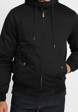 Homme portant une veste noire à capuche avec fermeture éclair à l'avant, cordons et deux poches à rabat, associée à un jean bleu foncé.