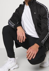 Chaquetón deportivo negro con acentos y logotipo blancos, combinado con pantalones ajustados negros y zapatillas blancas. Textura suave, cremalleras en los bolsillos.