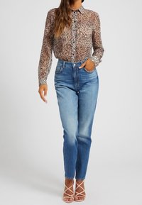 Kvinde iført en gennemsigtig, langærmet leopardmønstret bluse, der er stukket ind i højere talje, blå jeans og beige stropede hæle.