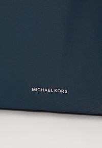 Tamsiai mėlynas nailono audinys; pasižymi elegantišku dizainu su matomu metaliniu "MICHAEL KORS" logotipu apačioje. Tekstūruota paviršius.