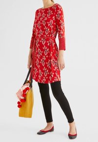 Punainen kukkakuvioinen mekko, jossa on kolme neljänneksen mittaiset hihat ja vyötärö istuvaksi nyörittynä; yhdistetty mustiin legginsseihin, punaisiin tasapohjaisiin kenkiin ja keltainen kangaskassi, jossa on kuvioitu huivi.