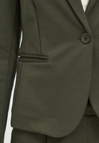 Dark green blazer with a smooth texture, featuring a single button closure and side pocket detail. Tailored fit and structured design.