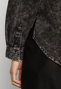 Camicia in denim nero con una texture sbiadita, dotata di polsini con bottoni e orlo curvo, mostrata su uno sfondo scuro.