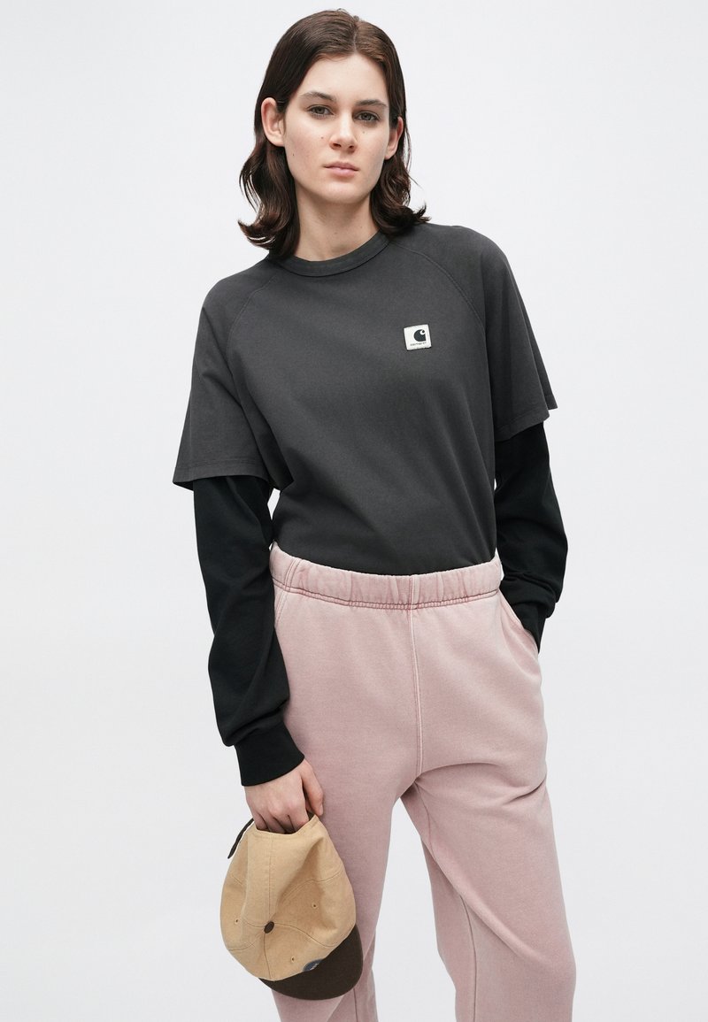 Giovane con capelli castani di media lunghezza che indossa una maglietta nera a maniche lunghe sovrapposta a una maglietta a maniche corte, pantaloni da tuta rosa, e tiene un cappellino beige.