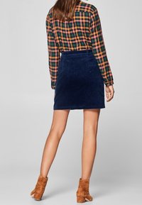 Jupe en velours côtelé navy avec une silhouette ajustée, associée à une chemise à carreaux multicolore. Le modèle porte des bottines beige.