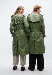 Πράσινο δερμάτινο παλτό τύπου trench με καφέ γιακά, ζώνη στη μέση και λεπτομέρεια σε πτερύγιο στην πλάτη. Φορώντας το δύο άτομα με σχέδια στα παντελόνια τους.