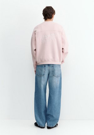 Persona che indossa una felpa rosa chiaro con testo bianco sulla schiena e jeans blu larghi, in piedi di fronte a un muro bianco.