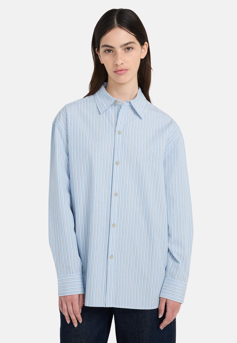 Νέα γυναίκα με ίσια σκούρα μαλλιά που φοράει ένα ανοιχτό γαλάζιο πουκάμισο μακρυμάνικο, φαρδύ, με κάθετες λευκές και καφέ ρίγες.