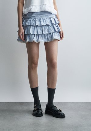 Osoba stojaca na sivom podlahe, oblečená v modro-bielej pásikavej vrstvenej mini sukni, bielom topu, tmavosivých ponožkách a čiernych kožených mokasínach.